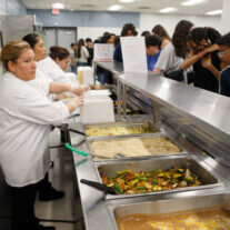 A cafeteria scene where several cafeteria staff in white uniforms are serving food to students lined up at the counter. Various dishes, including rice, vegetables, and sauces, are displayed in metal serving trays. The students, some holding trays, wait in line to be served.
