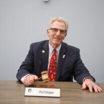 Phil Pritzker, wearing a dark suit and colorful tie, smiles while holding a gavel at a boardroom table, with a nameplate in front of him.