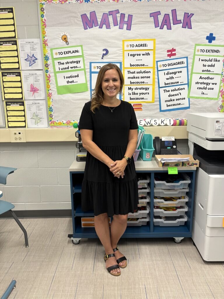 A woman in a black dress stands smiling in a classroom. The "Math Talk" board behind her features colorful posters with discussion prompts. The atmosphere is educational and inviting.