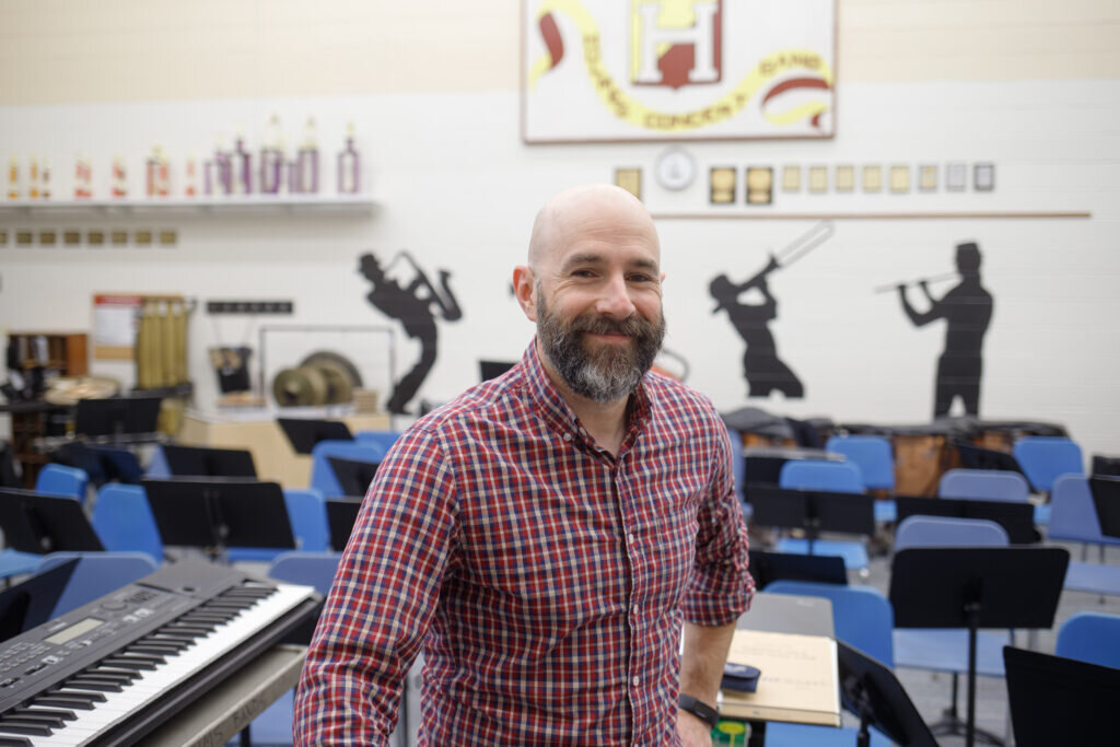 A smiling man in a red plaid shirt stands in a music classroom, surrounded by musical instruments and chairs. Wall silhouettes depict musicians playing.