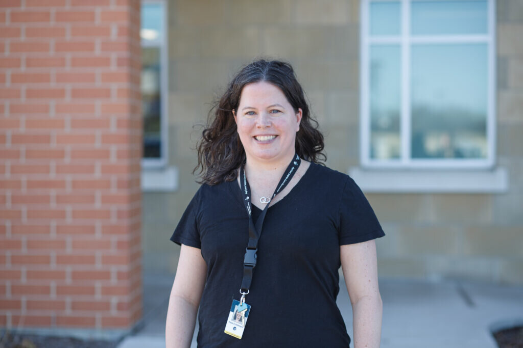 A woman with curly hair and a black shirt smiles warmly outside a brick building. She wears a lanyard with a badge. Background includes windows and a neutral wall.