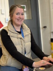 Smiling Megan Kenney in a beige vest works at a laptop in an office. A yellow travel mug and a blue lanyard are visible. The atmosphere is friendly.