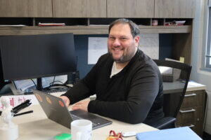Benjamin Neidich sits at his desk, smiling at the camera, with a laptop open. The office space features shelves with papers and a large monitor. The atmosphere is welcoming and professional.