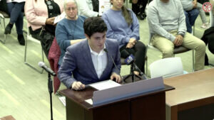 Ariel Lozner in a checked suit speaks at a podium with microphones, appearing focused. An audience sits attentively in a bright room.