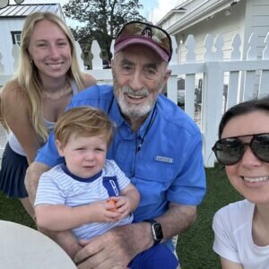 A family, including a smiling older man in a blue shirt (Jack Kimsey), a young child, and two women, pose outdoors by a white picket fence on a sunny day.