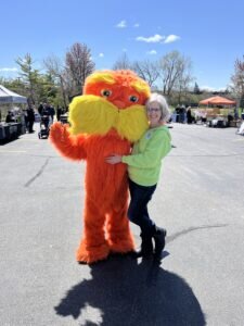 Mary Beth Schaye, in a green hoodie, hugs a person in a bright orange and yellow mascot costume, outdoors under a clear blue sky. It conveys a joyful, fun atmosphere.
