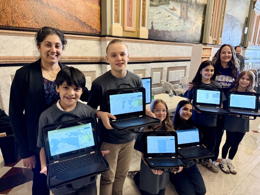 A group of smiling children and adults hold laptops displaying geographic maps, standing in a historic building with ornate paintings and marble walls.