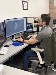 Anthony Mead. working on a computer with a 3D model displayed. He appears focused, with a tidy desk and a casual work environment visible.