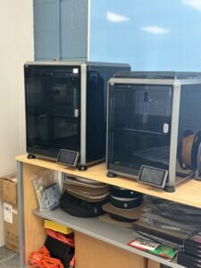 Two 3D printers on a shelf with accessories and supplies, including paper, scissors, and cartridges.