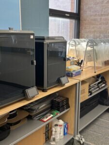 Two of D21's Creality 3D printers sit on a wooden shelf, surrounded by organized supplies and accessories. 