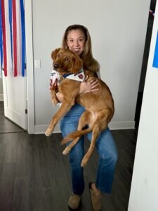Kayla Weiner smiling while holding a large brown dog (Mater) in a sports jersey, standing in a hallway with colorful streamers. The scene conveys a joyful mood.