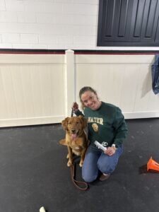 Kayla Weiner kneels beside her dog, Mater, holding his leash. She smiles at the camera, wearing a green sweater. Indoor setting with a white wall.