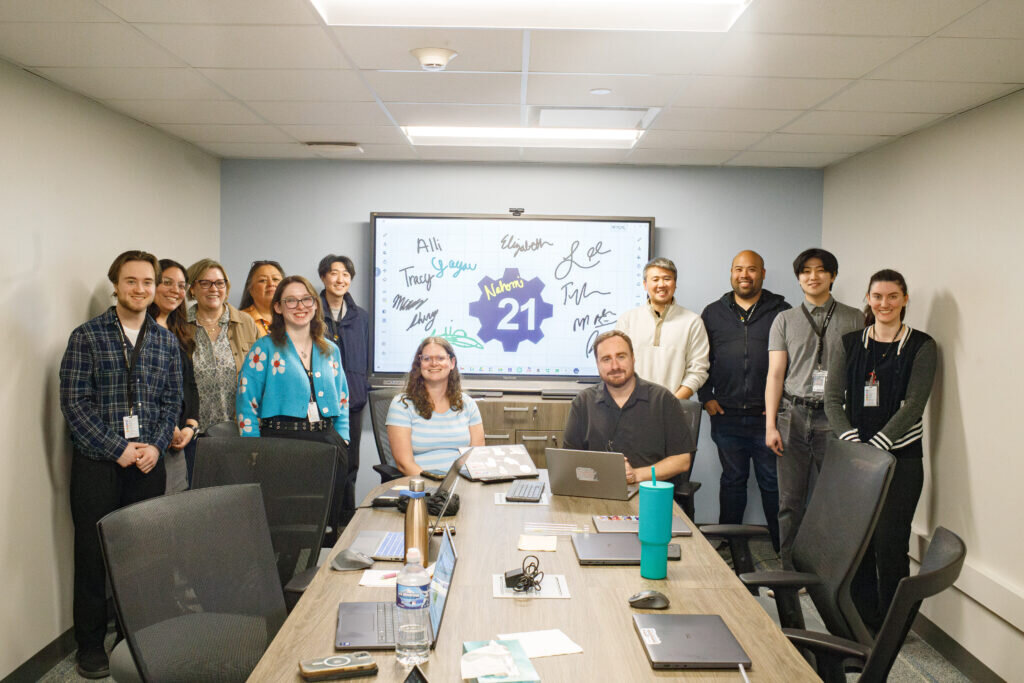 A group of twelve people, smiling, in a meeting room. A screen behind them shows "Welcome 21" with signatures. The atmosphere is friendly and collaborative.
