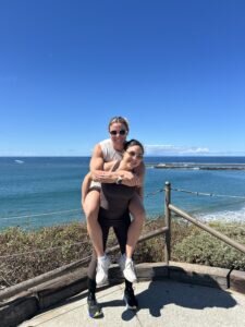 Kayla Weiner and Jenna Weiner smiling, with one giving the other a piggyback ride by the ocean. They are standing on a viewpoint with bright blue sky and sea.