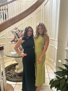 Kayla Weiner and Jenna Weiner in elegant dresses, one in dark green and the other in lime, stand smiling near a staircase. A floral arrangement is on a nearby table.