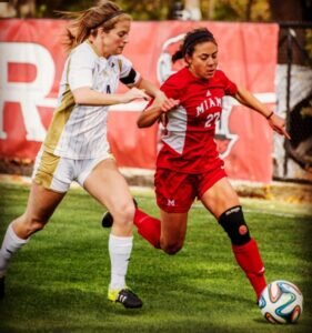 Jenna Weiner playing soccer, competing for the ball on a grassy field. She wears a red "Miami University" jersey, while the other wears a white kit. Intense action.