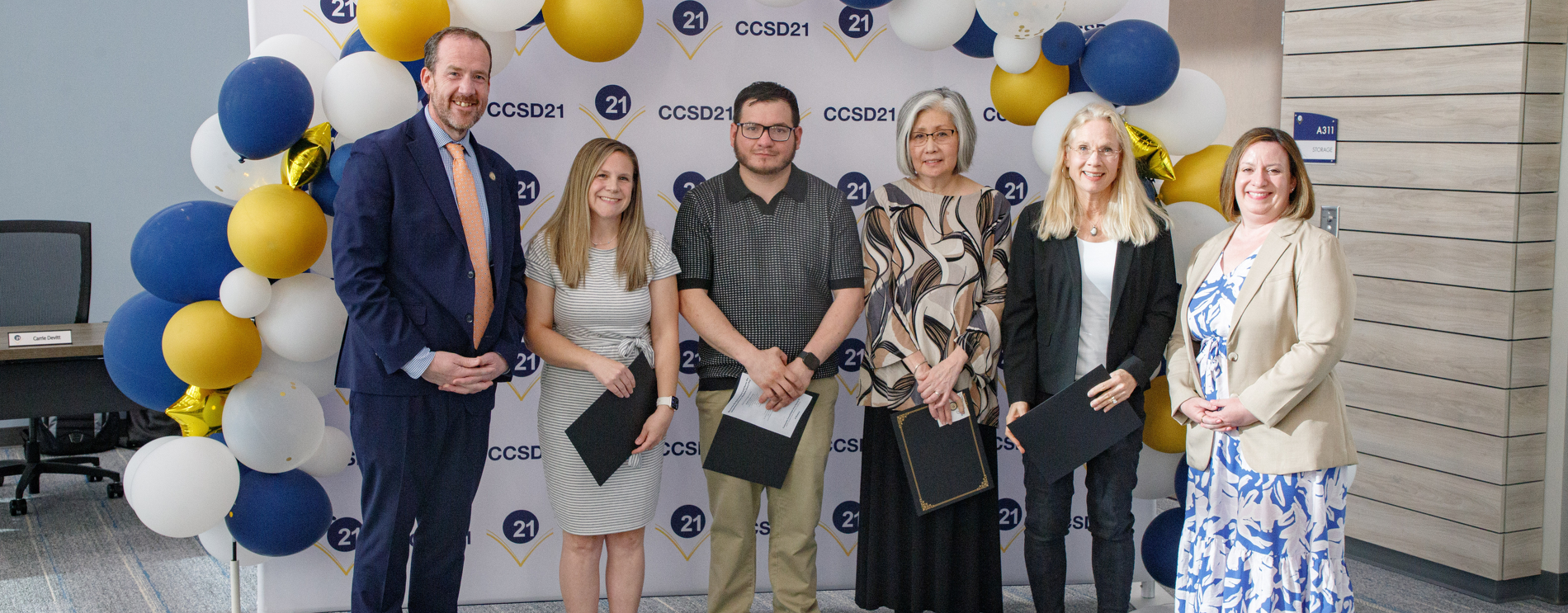 Six people stand in front of a blue and gold balloon arch and a CCSD21 backdrop, posing for a group photo at an indoor event.
