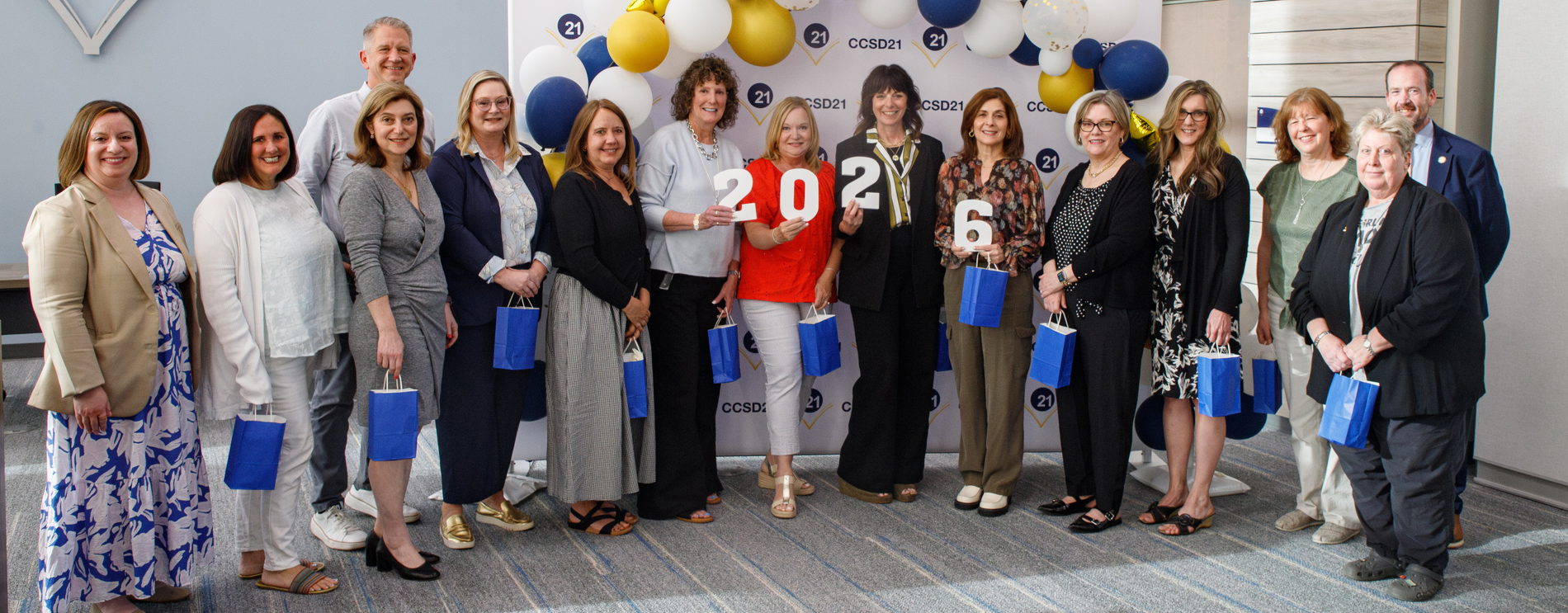 A group of people smiling, holding blue gift bags and numbers "2026." They're in front of a backdrop with CCSD21 branding and blue and yellow balloons. The atmosphere is celebratory.