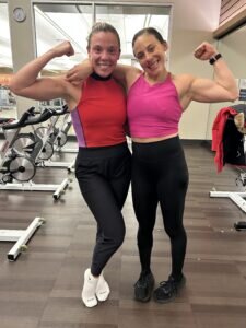 Jenna & Kayla Weiner pose in athletic with flexed arms, smiling in a gym. Exercise bikes are in the background.