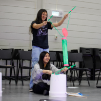 Two people engage in a creative indoor activity, using colorful materials and white buckets. They appear focused and amused, with chairs in the background.