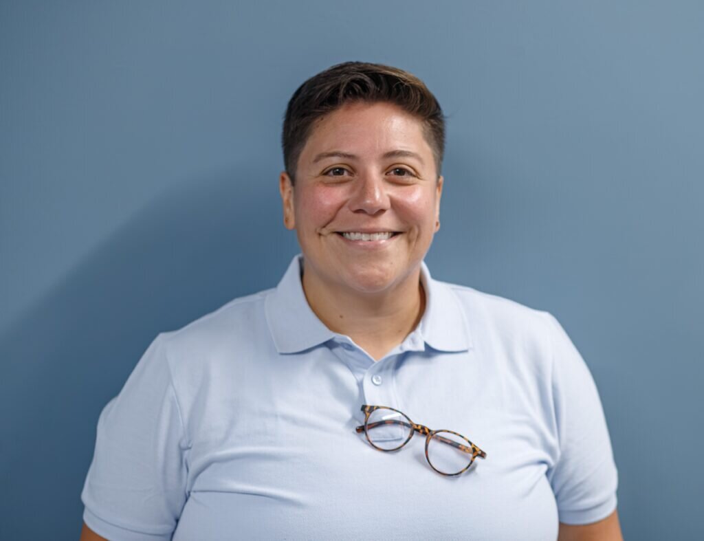 A medium close-up studio portrait of a person with short, dark hair styled to the side, smiling warmly. They are wearing a light blue polo shirt and have a pair of round, tortoiseshell-frame eyeglasses hanging from the shirt's button placket. The background is a solid, clean slate blue.