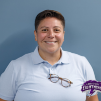 A medium close-up portrait of a smiling person with short, dark hair, wearing a light blue polo shirt against a solid blue background. A pair of tortoiseshell eyeglasses is hooked onto the collar of their shirt. In the bottom-right corner, there is a logo for "London Lightning" featuring purple text and a lightning bolt.